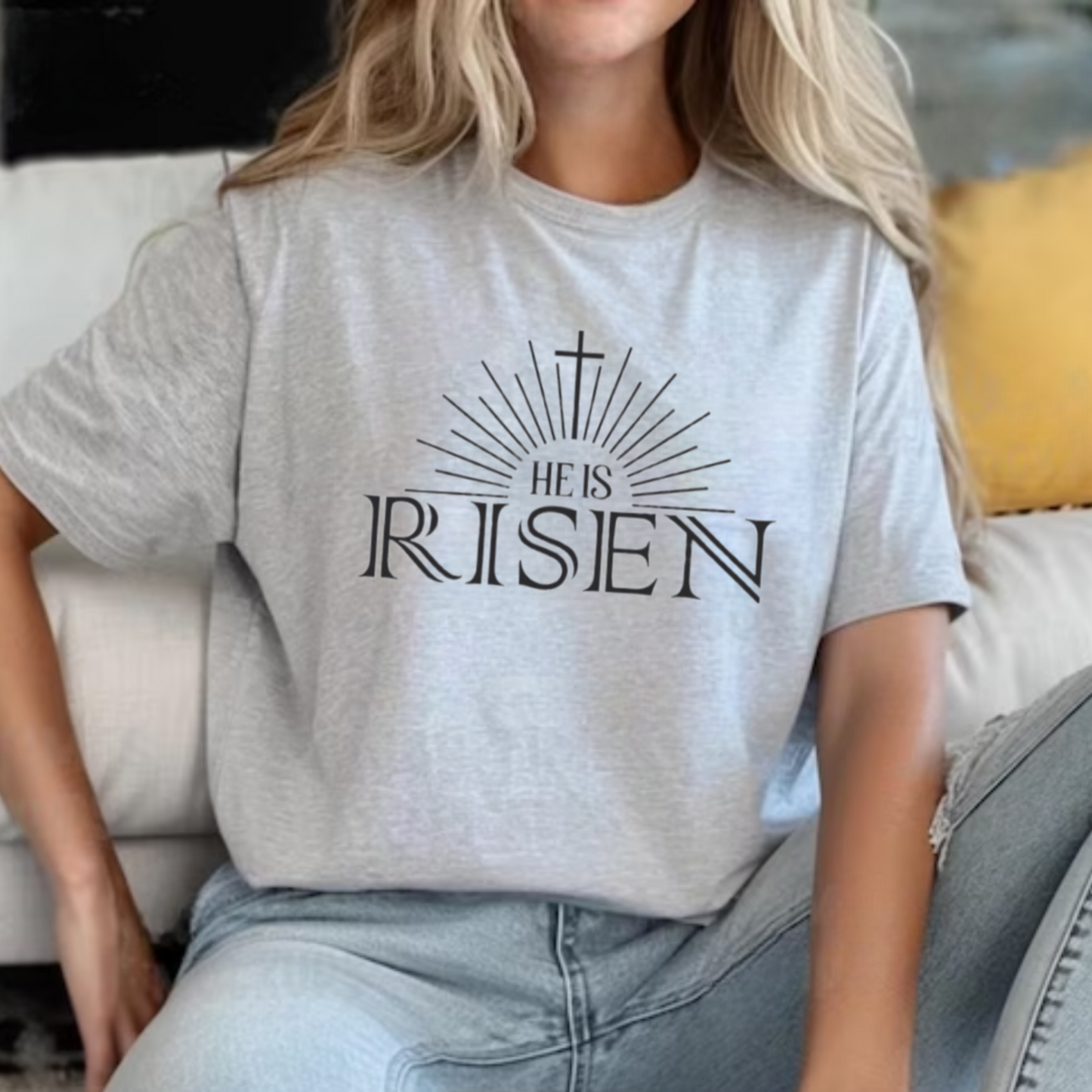 Woman sitting down wearing sports gray He Is Risen tee with cross sunburst graphic styled with ripped denim jeans with a couch in the background