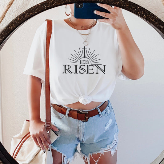 Woman wearing white He Is Risen tee with cross sunburst graphic styled with denim shorts and brown leather bag in round mirror selfie