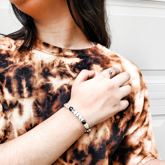 Close-up of Refined by Fire bleached tee showing unique black and tan tie-dye pattern detail with faith bracelet styling