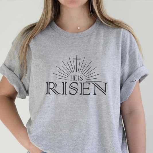 Woman wearing a sports gray He Is Risen tee with cross sunburst graphic styled with light wash jeans against a white background