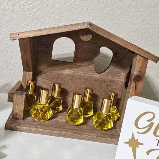 Nativity anointing oil blend in glass roller ball bottles with gold flakes are displayed in a wooden stable, with a white wall as the background.