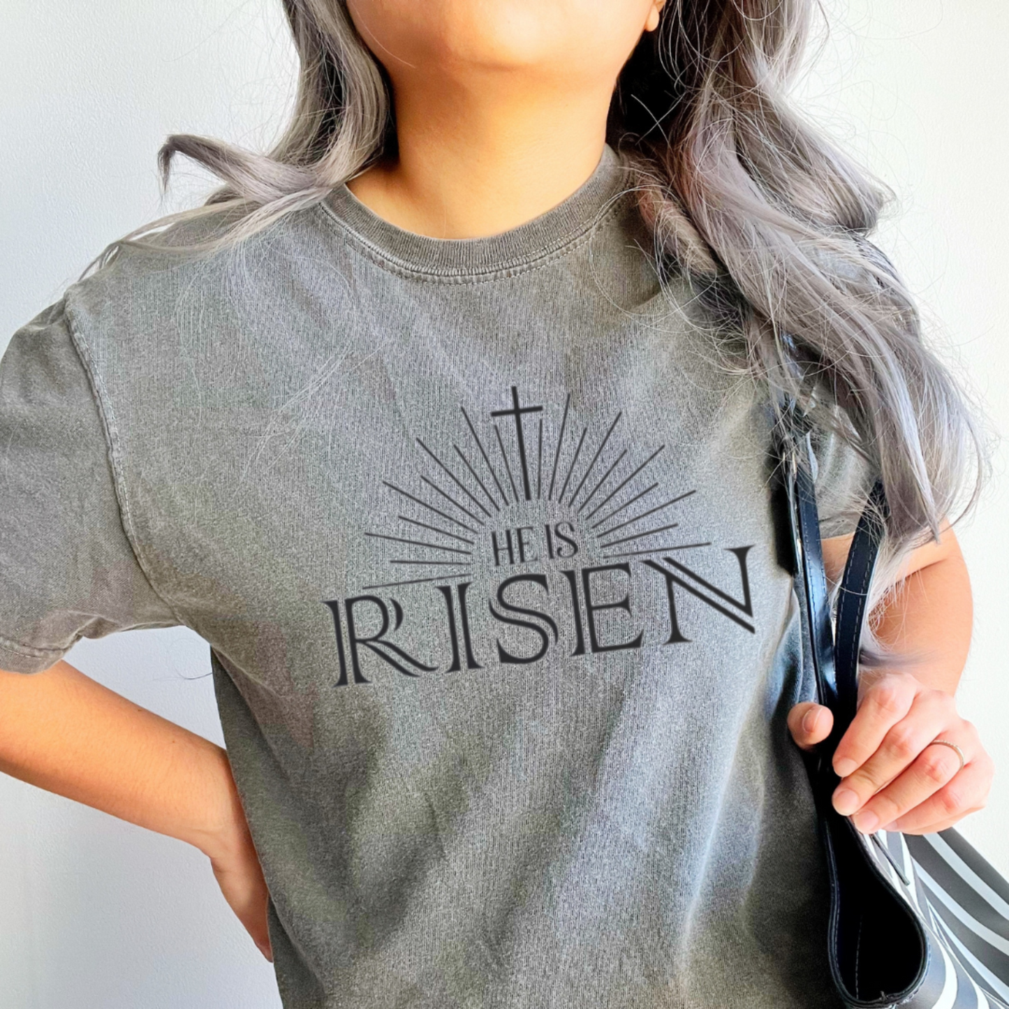 Woman wearing pepper colored He Is Risen tee with cross sunburst graphic styled with light wash jeans and striped bag