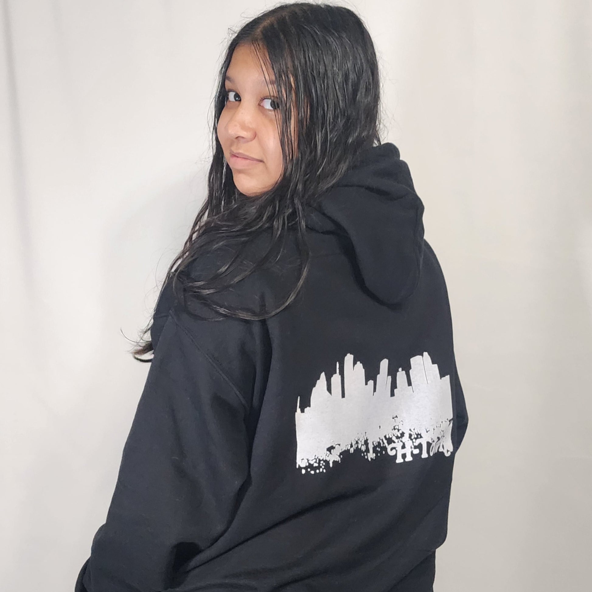 Teen turning around in front of a white background to show the back of a black I've Witnessed It graphic hoodie showing the Houston Skyline