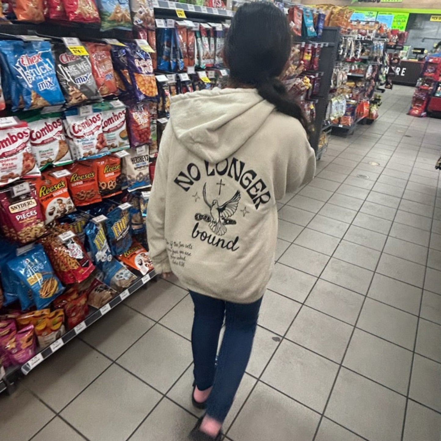 Personwearing an ivory No Longer Bound hoodie while walking through a grocery store aisle with snacks on shelves.