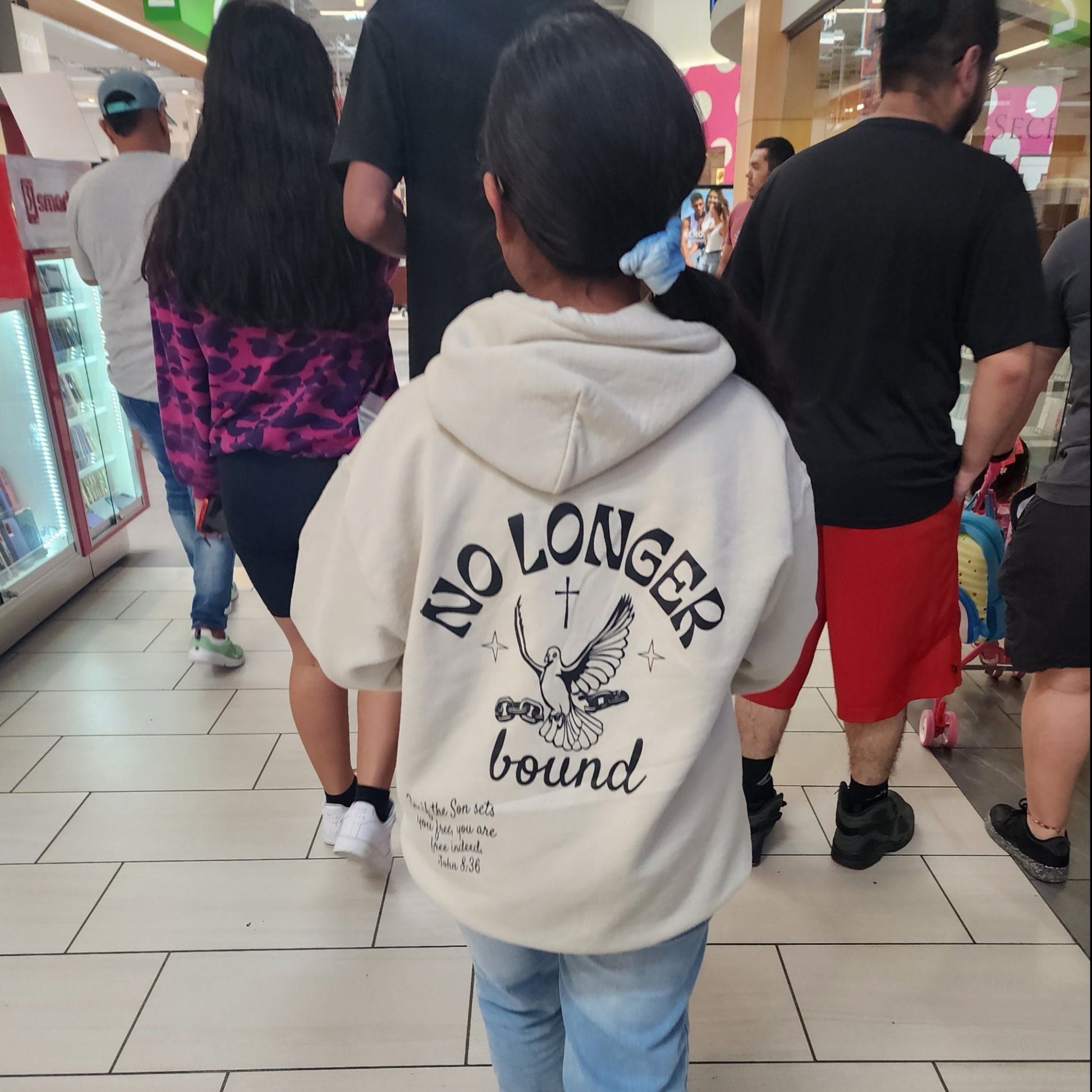 Woman wearing a n ivory No Longer Bound hoodie while walking thorough a shopping mall