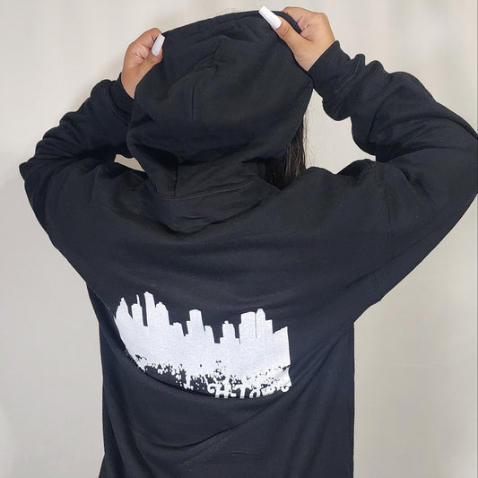 Teen in front of a white background showing off the back of a black I've Witnessed It graphic hoodie showing the Houston Skyline 